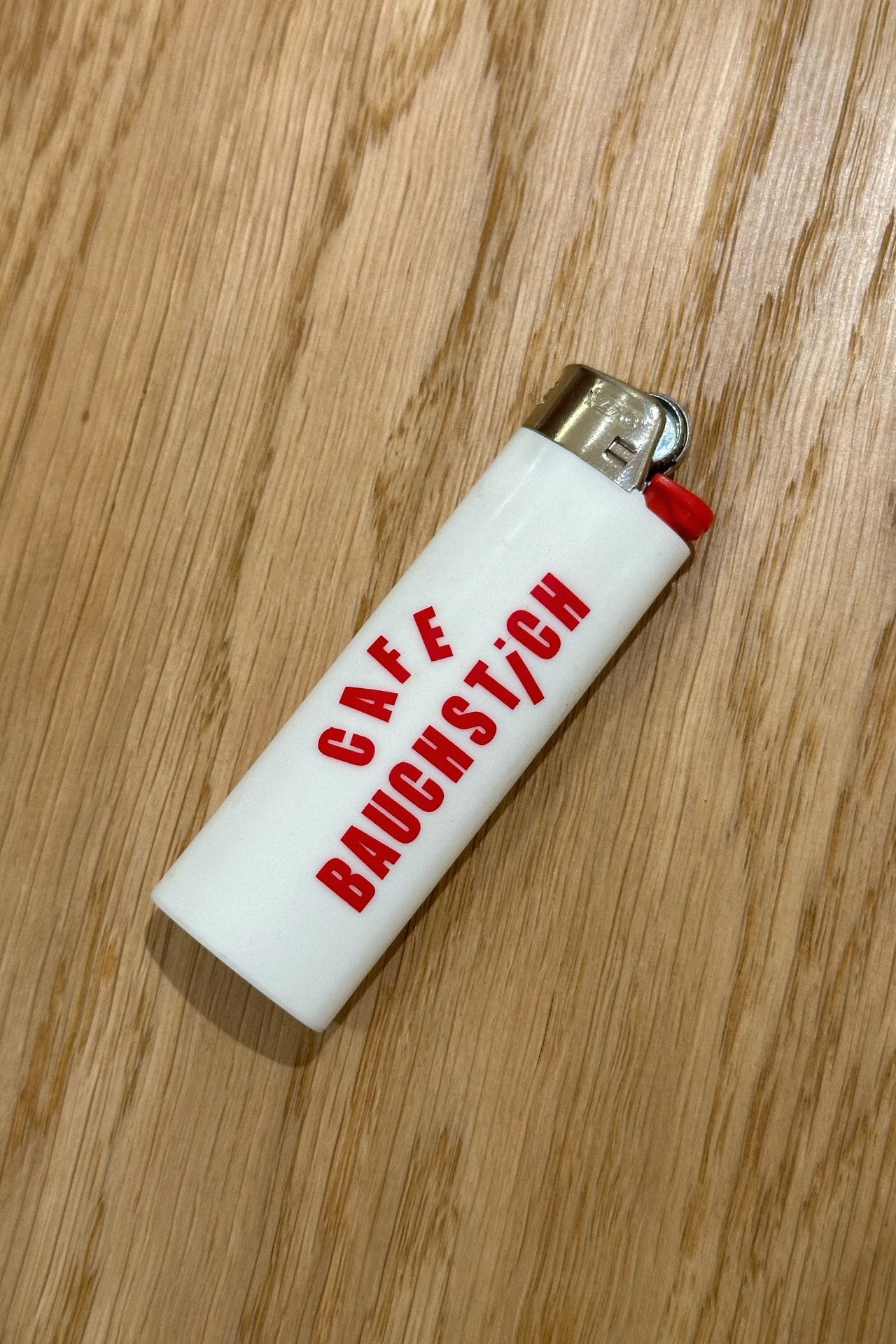 Weißes BIC Feuerzeug mit rotem Cafe Bauchstich Logo, Wien Streetwear, Vorderseite auf Holzuntergrund